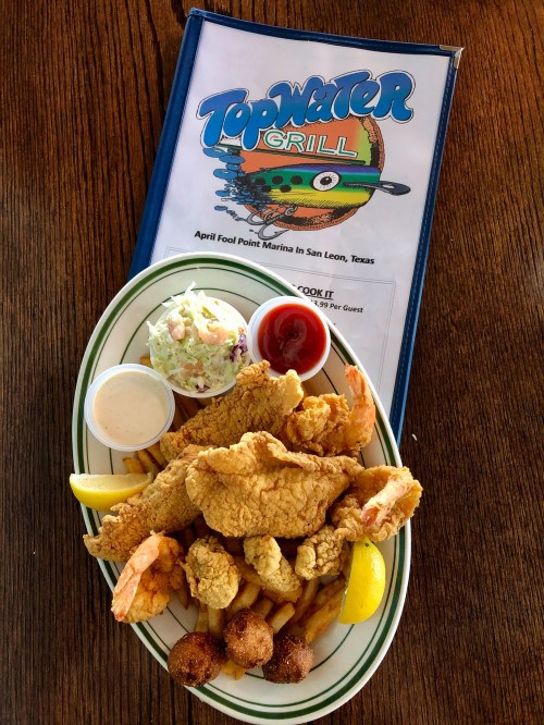 A plate of fried seafood including shrimp, fish, and hushpuppies, served with coleslaw and dipping sauces, next to a menu.