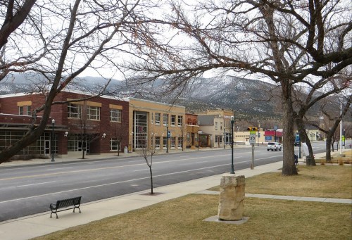 A quiet street lined with buildings, bare trees, and mountains in the background on a cloudy day.