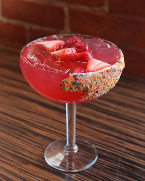 A vibrant pink cocktail in a glass, garnished with fresh strawberries and a colorful, sugary rim.