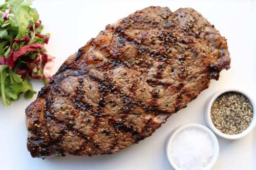 Grilled steak with a charred surface, accompanied by fresh greens and bowls of salt and pepper.