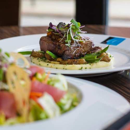 A plated dish featuring a steak on creamy mashed potatoes, garnished with microgreens and a side salad.