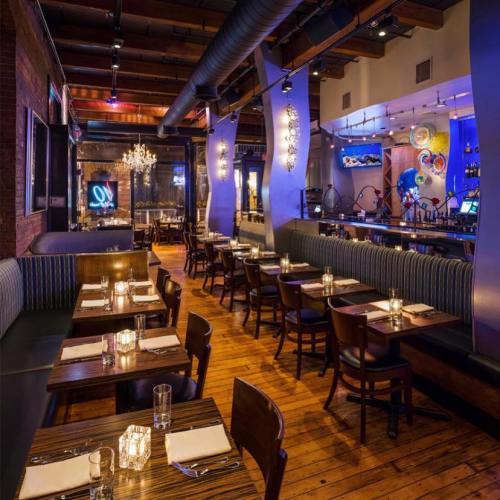 A stylish restaurant interior with wooden floors, dim lighting, and a bar in the background, featuring elegant decor.