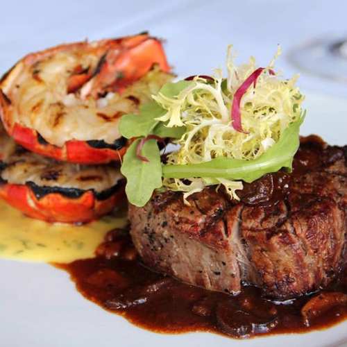 A plated dish featuring grilled shrimp, a beef steak with sauce, and garnished greens on a white background.