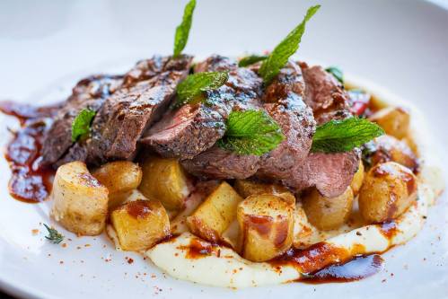 A plate of sliced beef with mint leaves, served on a bed of creamy mashed potatoes and roasted potatoes, drizzled with sauce.