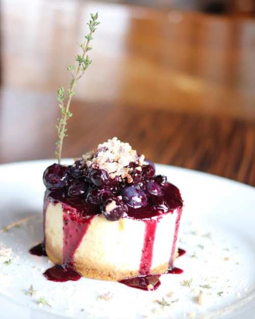 A slice of cheesecake topped with blueberry sauce, crushed nuts, and a sprig of thyme on a white plate.