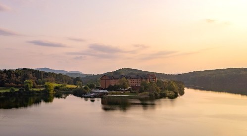 A serene landscape featuring a calm lake, lush greenery, and a distant hotel surrounded by mountains at sunset.