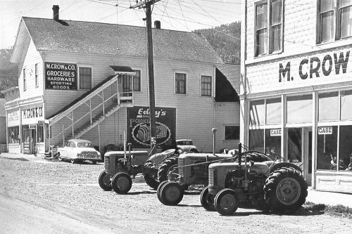 Black and white photo of a street scene featuring vintage tractors, a hardware store, and a parked car.