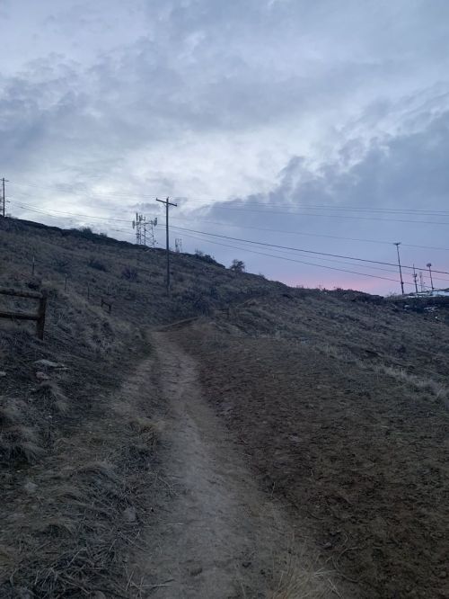 A winding dirt path leads up a hillside under a cloudy sky, with power lines in the background.