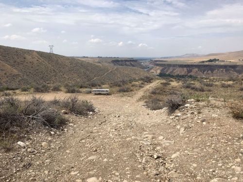 A rocky dirt path leads through a dry landscape with sparse vegetation, overlooking a distant canyon under a cloudy sky.