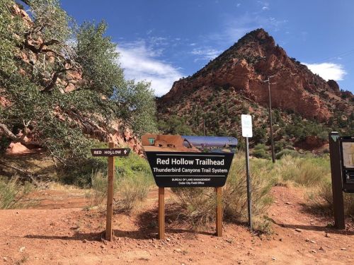 Sign for Red Hollow Trailhead in a desert landscape, with red rock formations and blue sky in the background.