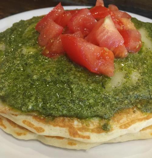 A stack of pancakes topped with green pesto and diced tomatoes on a white plate.