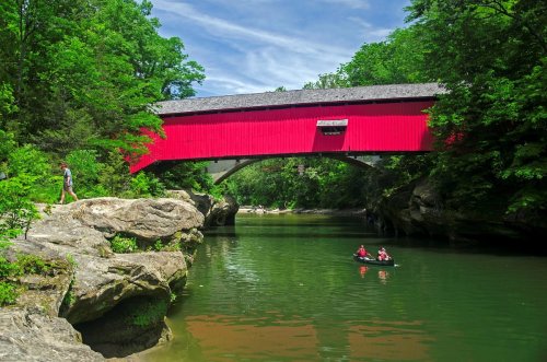 A red covered bridge spans a serene river surrounded by lush greenery, with people kayaking nearby.
