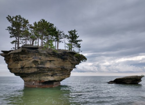 A rocky island with trees on top, surrounded by calm water under a cloudy sky.