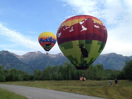 Two colorful hot air balloons float above a grassy field with mountains in the background and people watching nearby.