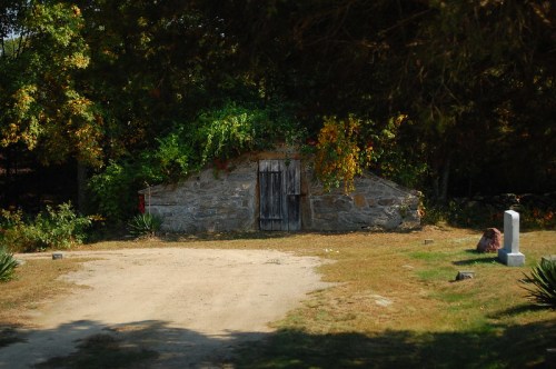 A stone structure with a wooden door, surrounded by greenery, set in a grassy area with a few scattered stones.