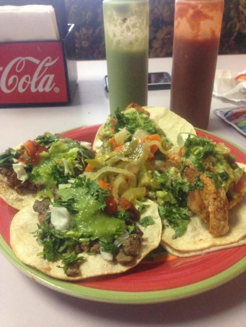 Three tacos on a colorful plate, topped with meat, fresh herbs, and sauces, with drinks in the background.