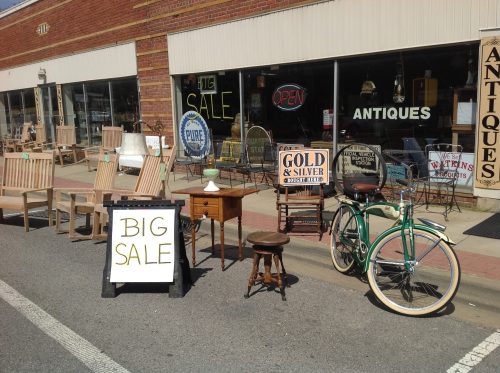 A storefront with various antique furniture for sale, including chairs, tables, and a bicycle, with a "Big Sale" sign.