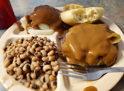 A plate of food featuring a meat patty with gravy, mashed potatoes, black-eyed peas, and a bread roll.