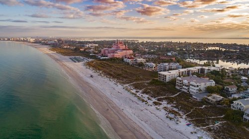 Aerial view of a beach with a pink hotel, coastal homes, and a sunset sky over calm waters.