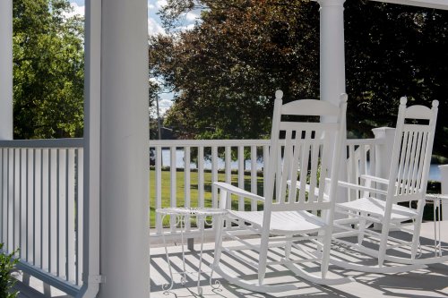 Two white rocking chairs on a porch overlooking a green lawn and a body of water, framed by trees and blue sky.
