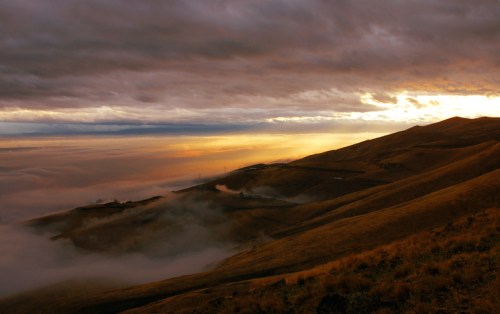 A dramatic landscape at sunset, with rolling hills shrouded in mist and a colorful sky filled with clouds.
