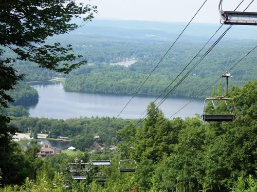 A scenic view from a ski lift overlooking a lake surrounded by lush green trees and distant hills.