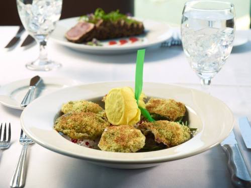 A plate of baked oysters with a yellow garnish, served on a bed of greens, with a glass of water in the background.