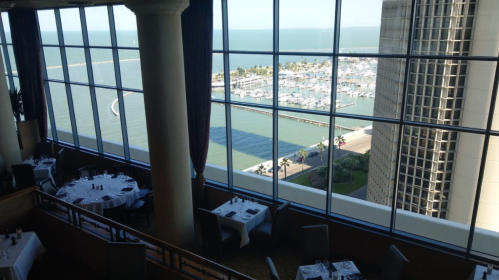 View from a restaurant overlooking a marina with boats and the ocean, featuring elegant dining tables in the foreground.