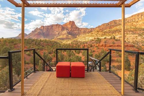 A scenic view from a wooden deck with two red cushions, overlooking mountains and lush greenery under a blue sky.