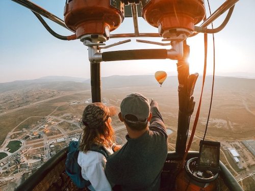 A couple in a hot air balloon, pointing at another balloon in the distance during sunset over a scenic landscape.