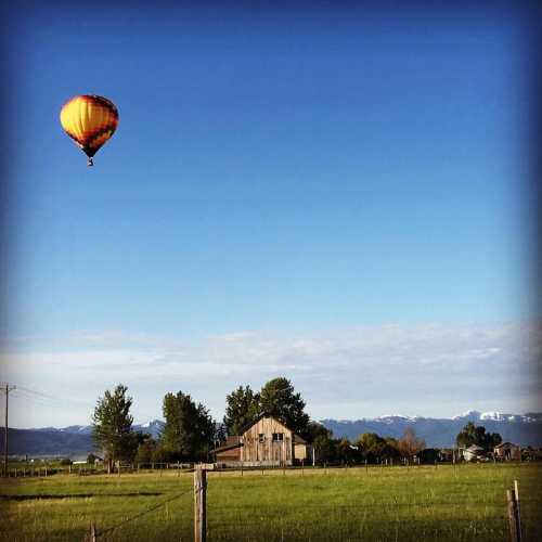 A colorful hot air balloon floats above a rural landscape with a barn and mountains in the background under a clear blue sky.