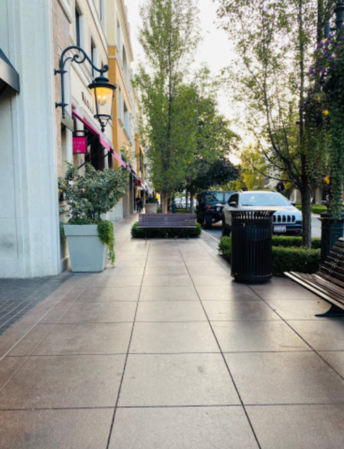 A tree-lined sidewalk with benches, planters, and parked cars, illuminated by street lamps in a pleasant urban setting.