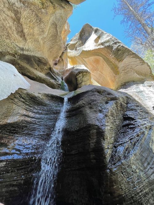 A waterfall cascades down smooth, rocky cliffs under a clear blue sky, surrounded by towering stone formations.