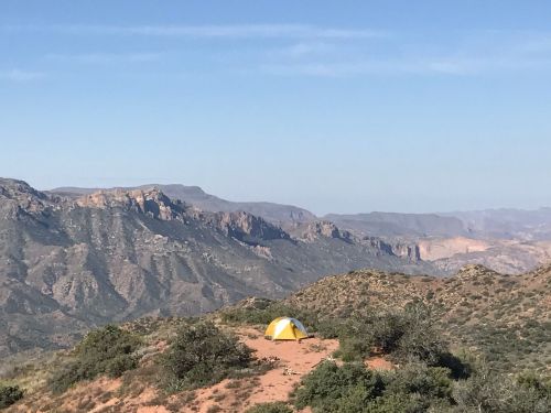 A yellow tent set on a sandy area, surrounded by mountains and a clear blue sky in the background.