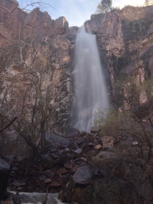 A tall waterfall cascades down rocky cliffs, surrounded by sparse trees and boulders in a natural setting.
