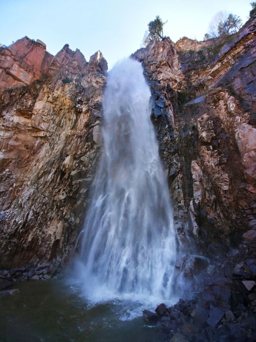 A powerful waterfall cascading down rocky cliffs into a serene pool below, surrounded by rugged terrain.