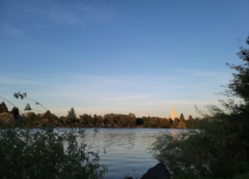 A serene lake scene at dusk, with trees lining the shore and a distant building illuminated by the setting sun.