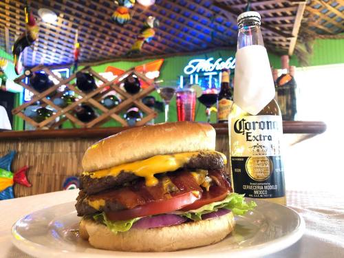 A juicy cheeseburger with lettuce, tomato, and onion, alongside a bottle of Corona beer in a colorful restaurant setting.
