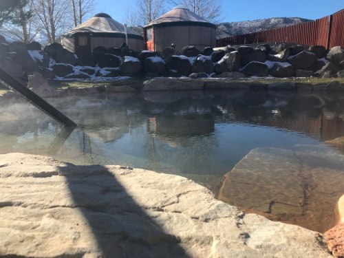 A serene hot spring surrounded by rocks and yurts, with steam rising in a tranquil outdoor setting.