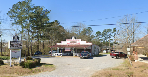 A small roadside restaurant with a sign, surrounded by trees and parked cars on a gravel lot.