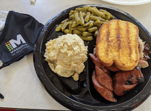 A black plate with grilled sausage, green beans, mashed potatoes, and toasted bread, alongside a branded face mask.