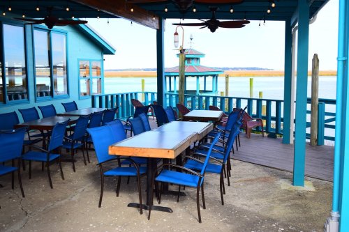 A waterfront restaurant patio with blue chairs and tables, overlooking a calm waterway and a small gazebo.
