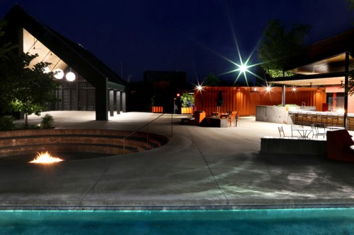 A modern outdoor space at night featuring a pool, fire pit, and stylish seating areas surrounded by greenery.