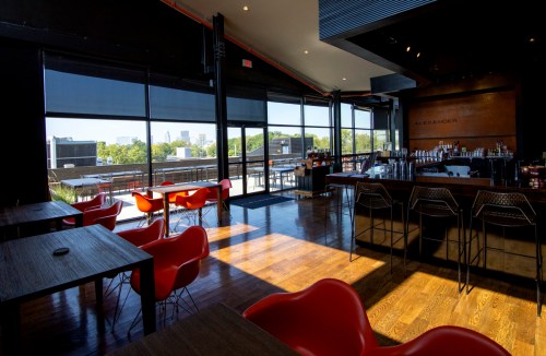 Modern bar interior with wooden floors, red chairs, and large windows showcasing a city view.