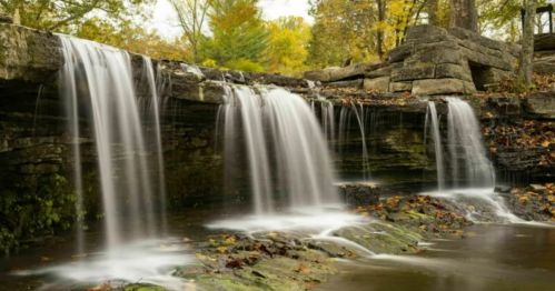A serene waterfall cascades over rocky ledges, surrounded by autumn foliage and a tranquil pool below.