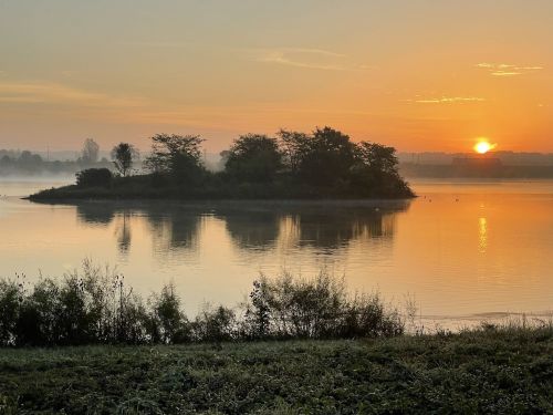 A serene sunrise over a calm lake, with an island and misty trees reflected in the water.