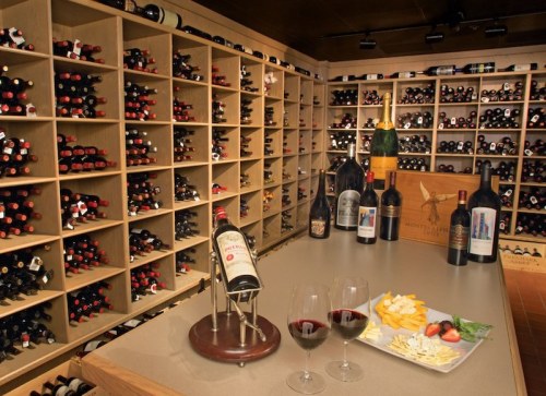 A wine cellar with shelves of bottles, a tasting table with two glasses of red wine, cheese, and strawberries.
