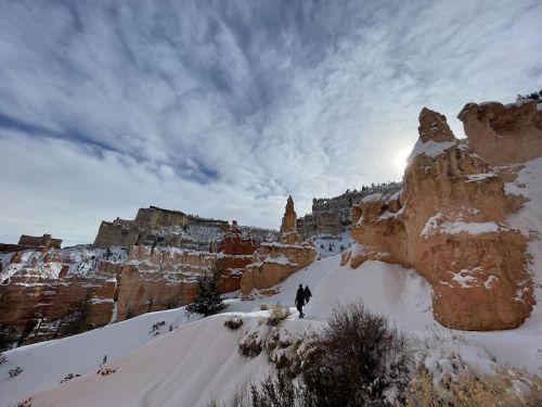 A person walks through a snowy canyon landscape with towering rock formations and a cloudy sky.