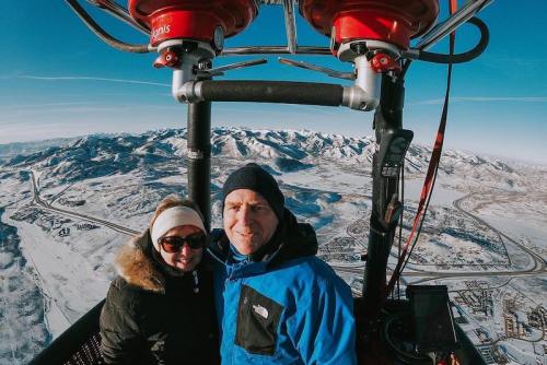 A couple smiles in a hot air balloon, overlooking snowy mountains and a scenic landscape below.