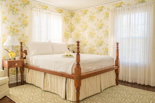 Cozy bedroom with floral wallpaper, a four-poster bed, and soft natural light from large windows.